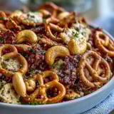 Golden-brown Everything Ranch Cheese and Pretzel Snack Mix spills from a white bowl, featuring crunchy pretzels, cheesy crackers, and cashews glistening with savory seasoning.  