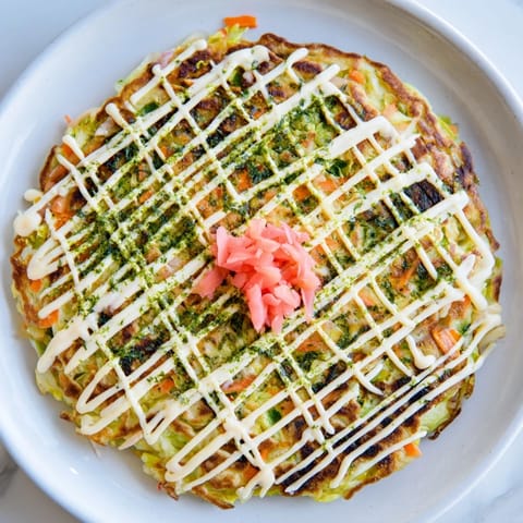 Fluffy Japanese Okonomiyaki Pancakes layered with shredded cabbage, bacon, and green onions, finished with savory sauce and vibrant pickled ginger garnish.